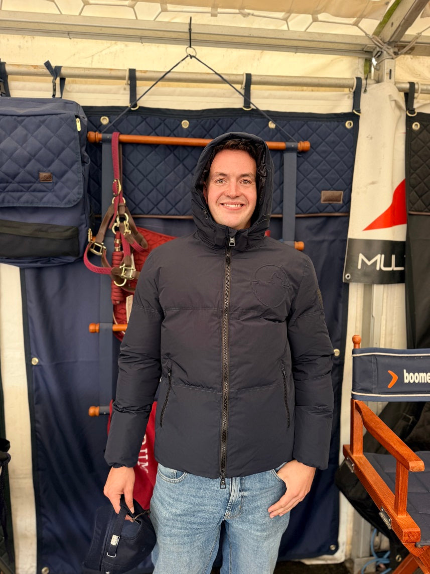 A man wearing a black hooded puffer jacket and blue jeans stands inside a tent-like stall with navy quilted tack or storage bags hanging behind him.