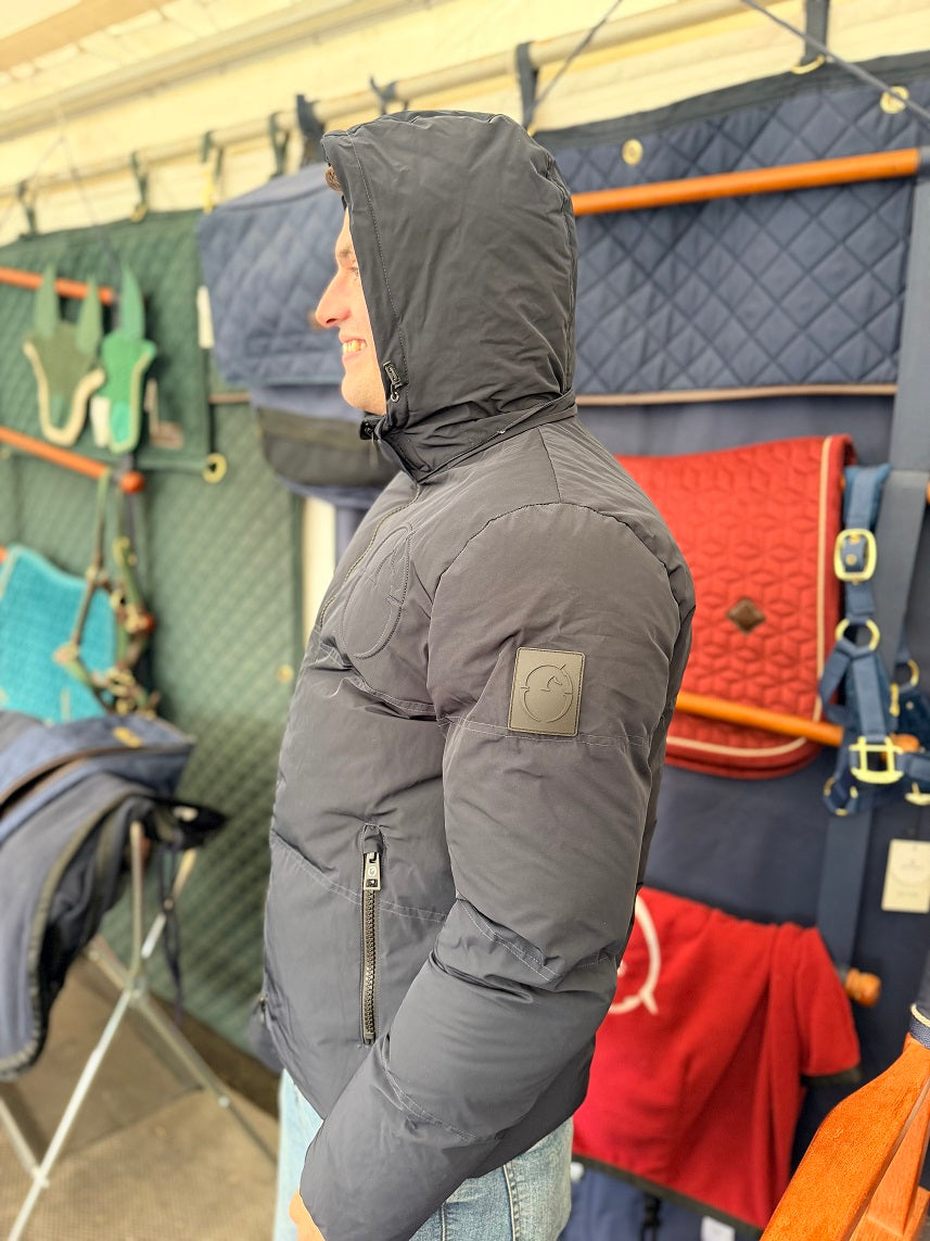 A man wearing a dark gray hooded puffer jacket with a zippered chest pocket, standing in a retail store next to a display of saddles.
