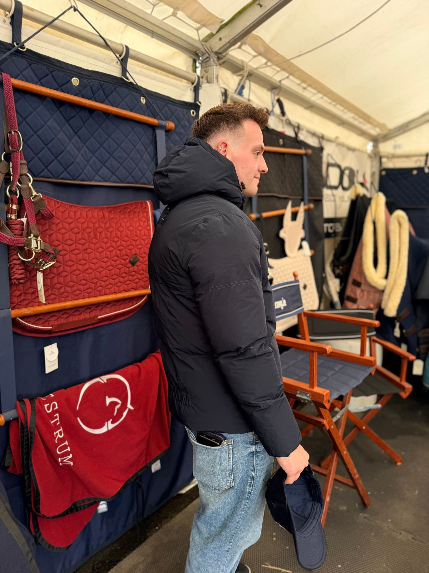 A man in a black puffer jacket and blue jeans stands inside a tent-like stall, looking at horse tack and saddlery items hanging on display. Visible products include a red quilted horse bag, red and navy blankets with white branding, and wooden folding chairs.