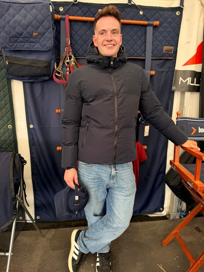 A man wearing a dark gray puffer jacket, blue jeans, and black and white sneakers stands in front of a navy blue quilted storage backdrop, holding a navy cap.