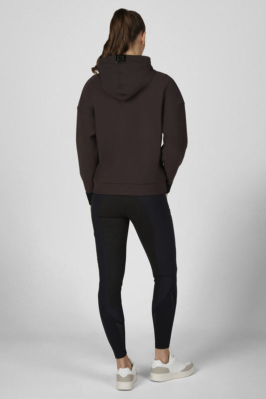 A woman is shown from the back wearing a dark brown pullover hoodie and black leggings, paired with white sneakers, against a light gray studio background.