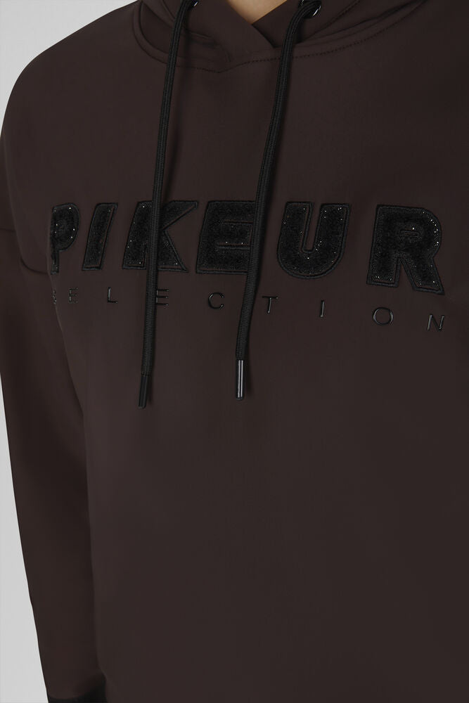 A close-up of a dark brown hoodie featuring a black glittery 'PIKEUR' logo and 'SELECTION' text across the chest, with black drawstrings.