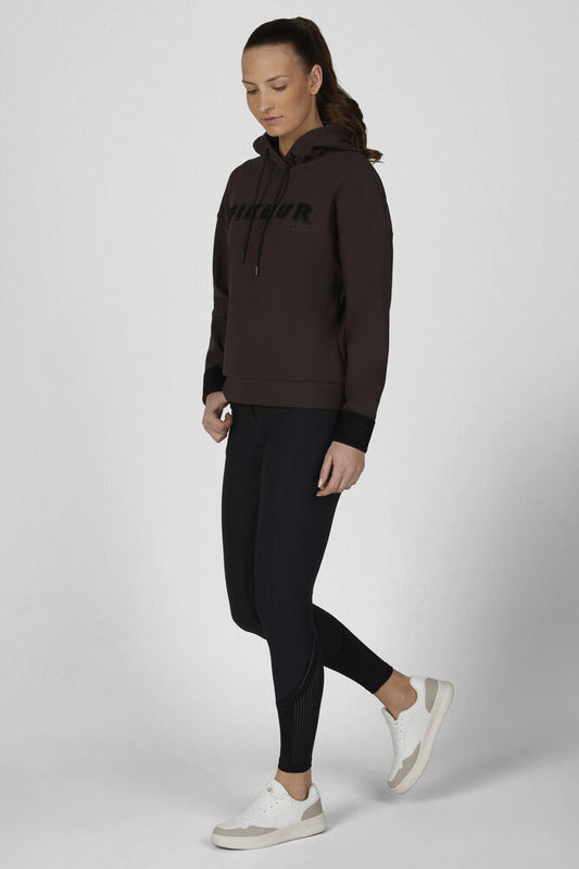 A woman modeling a dark brown pullover hoodie with black drawstrings and black 'HIBUR' text on the chest, paired with black leggings and white sneakers, against a light gray studio background.