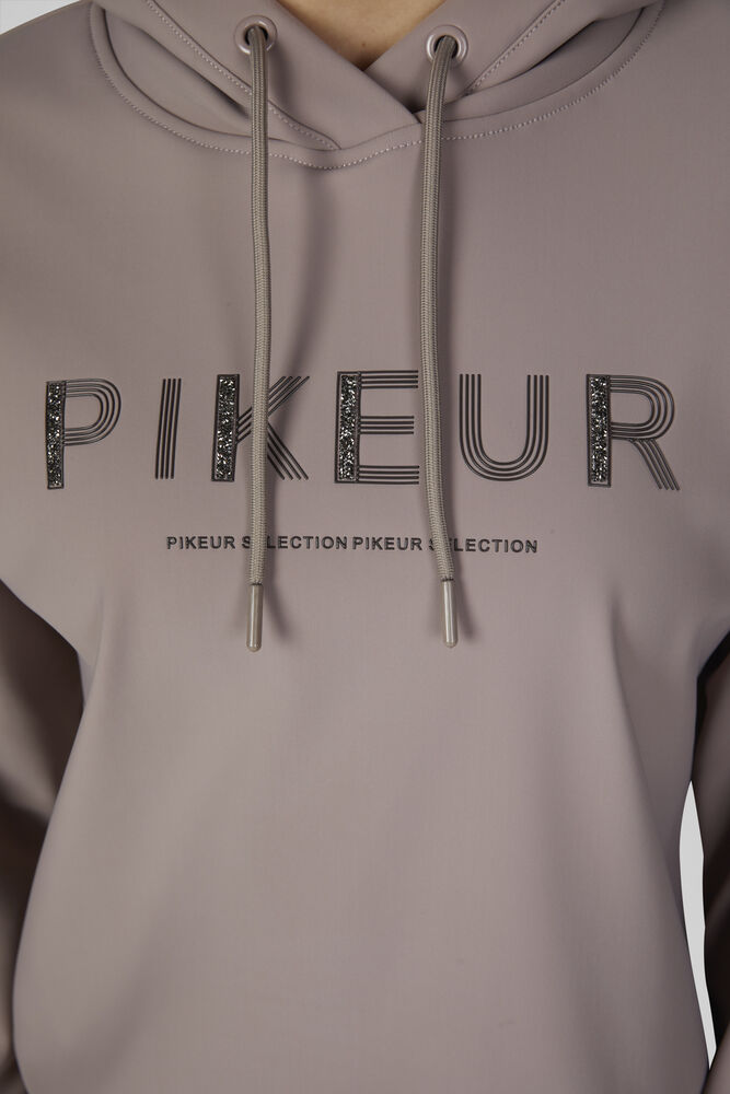 A close-up of a person wearing a muted taupe-brown pullover hoodie with 'PIKEUR' printed across the chest in black outlined lettering with glittery fill, and smaller 'PIKEUR SELECTION' text below. The hoodie has matching drawstrings and is shown against a plain light background.
