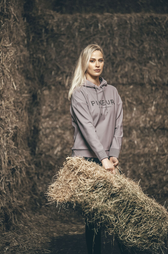 A blonde woman wearing a taupe-gray Pikeur hoodie stands in a barn holding a hay bale. The hoodie features the brand name 'PIKEUR' printed in black across the chest.
