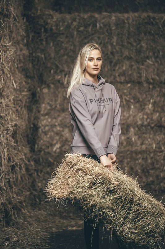 A blonde woman wearing a taupe-gray Pikeur hoodie stands in a barn holding a hay bale. The hoodie features the brand name 'PIKEUR' printed in black across the chest.