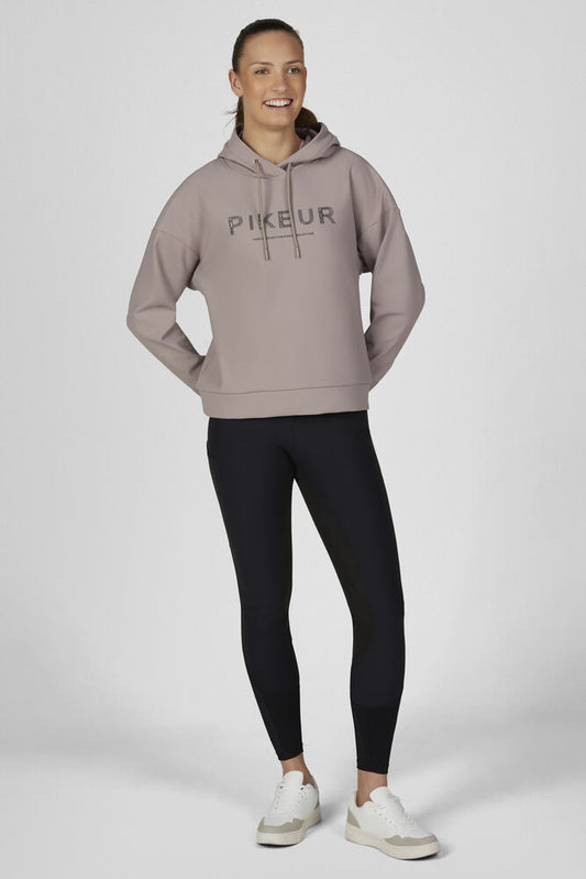 A woman modeling a light taupe Pikeur pullover hoodie with black leggings and white sneakers, standing against a neutral light gray studio background.