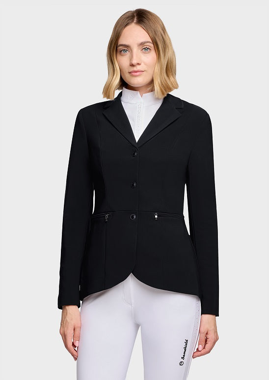 A woman modeling a black equestrian riding jacket over a white mock neck shirt and white riding breeches, photographed against a light gray studio background.