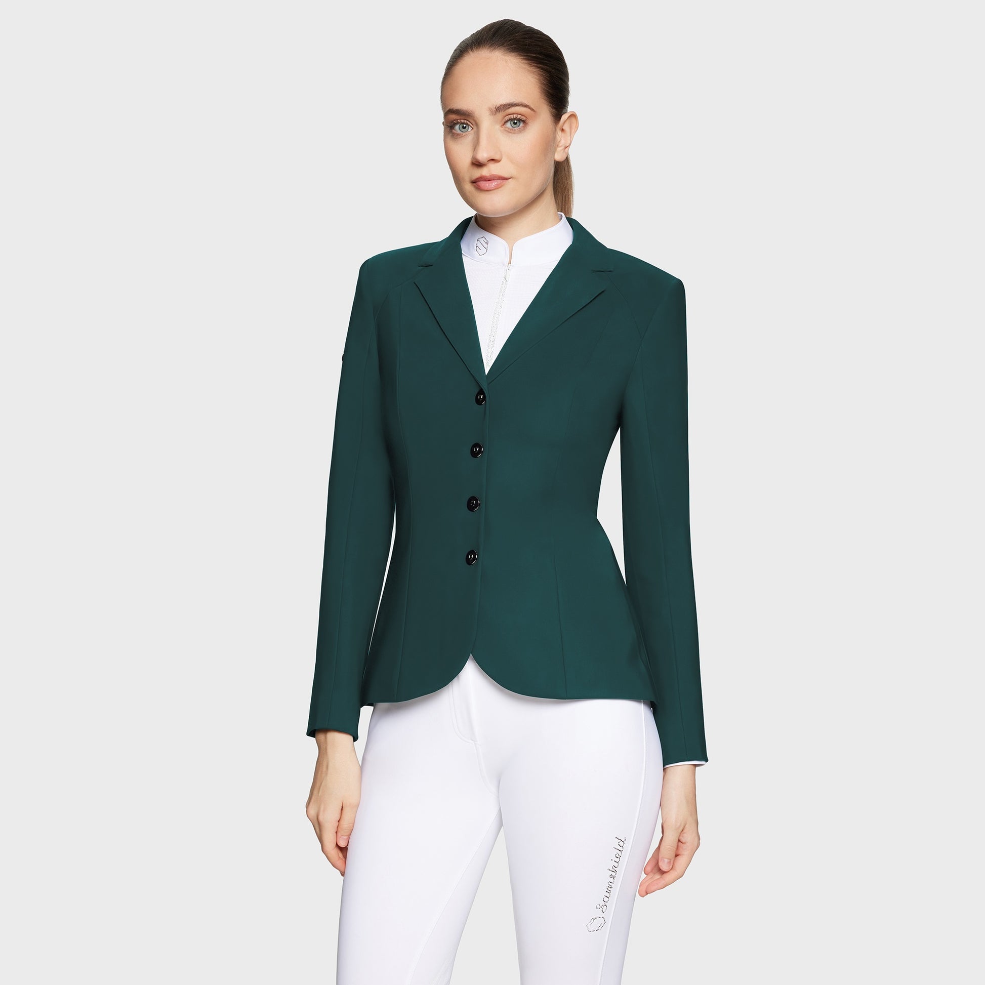 Woman wearing a green riding jacket and white pants on a light gray background