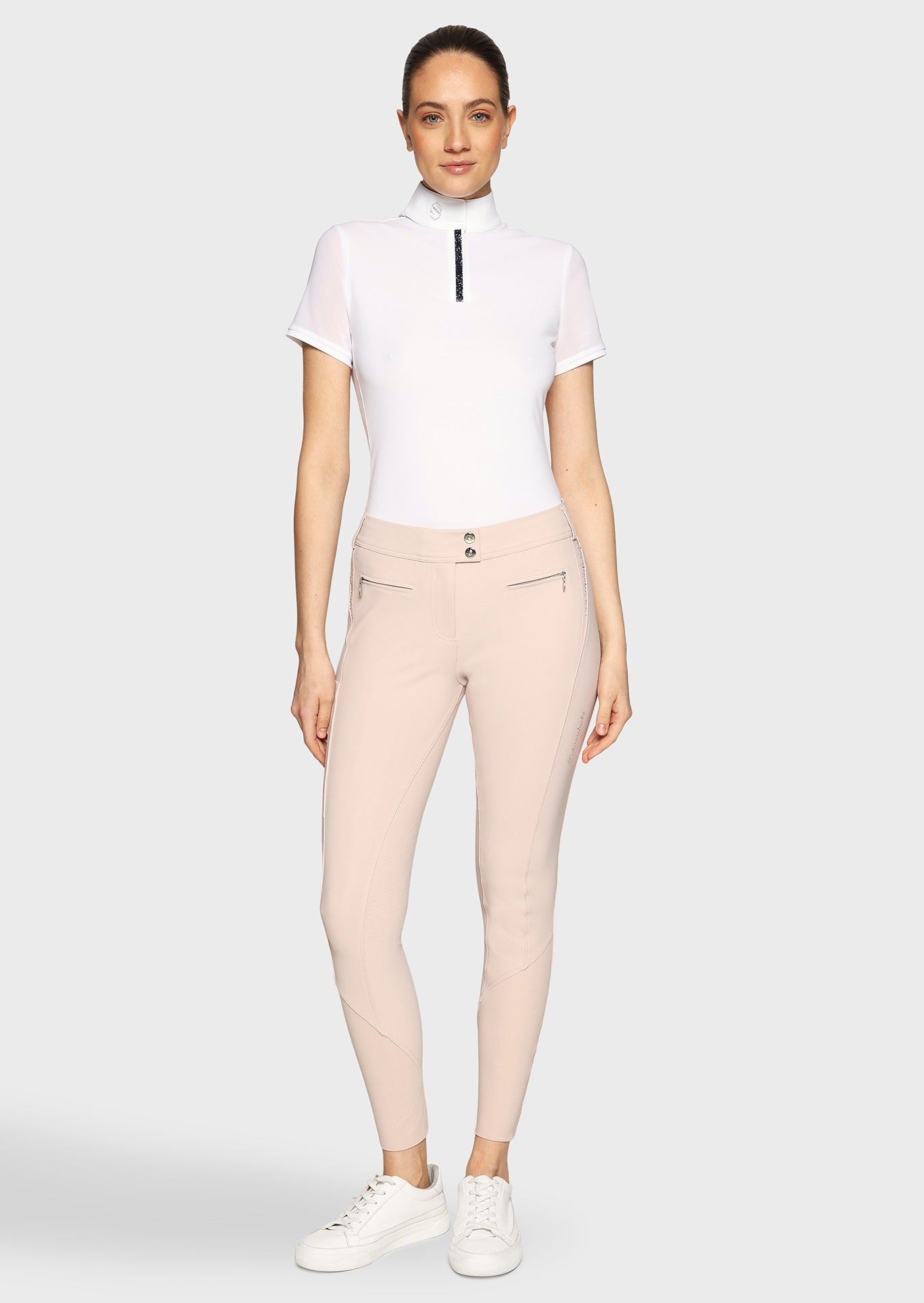 Woman wearing a white competition shirt and pink breeches on a beige background