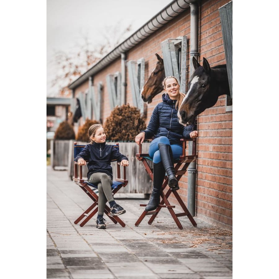 Two individuals sitting on foldable director's chairs outdoors, with horses peeking out from behind a wall.