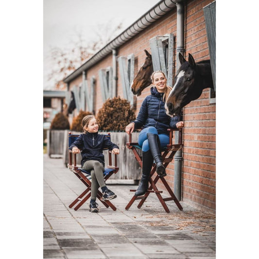 Two individuals sitting on foldable director's chairs outdoors, with horses peeking out from behind a wall.