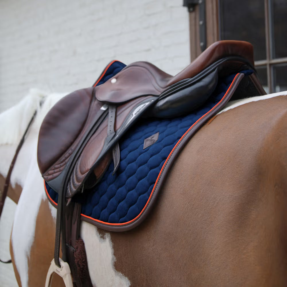 Brown saddle on a horse with a blue pad against a white brick wall.