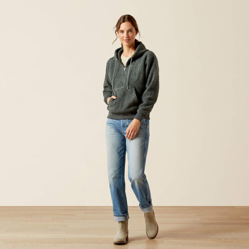 Woman wearing a dark green hoodie, light blue jeans, and beige boots on a plain background
