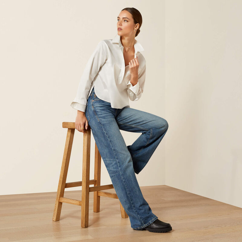 Woman leaning on a stool wearing a white shirt and blue jeans against a beige background