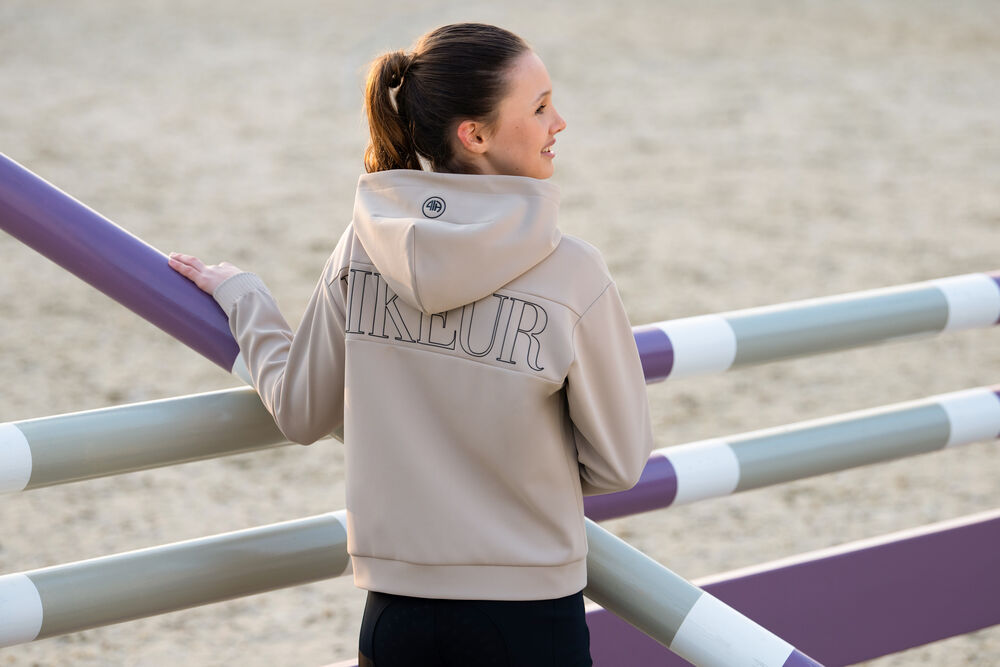 Person wearing a beige hoodie with 'Pikeur' branding, standing near equestrian jumps.