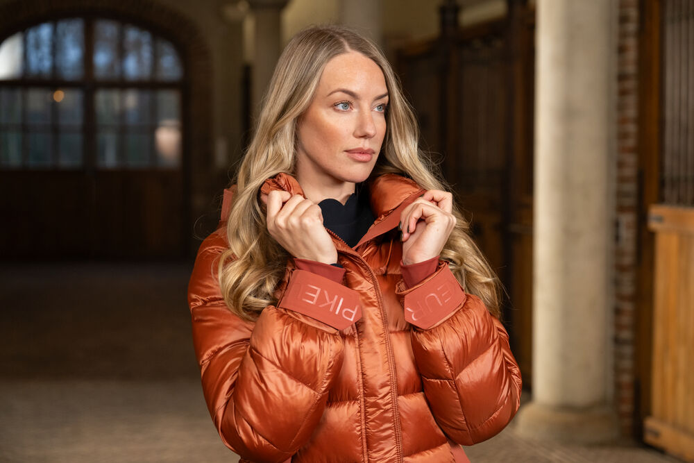 Woman wearing a brown puffer jacket with 'PIKEUR' branding in an indoor setting.