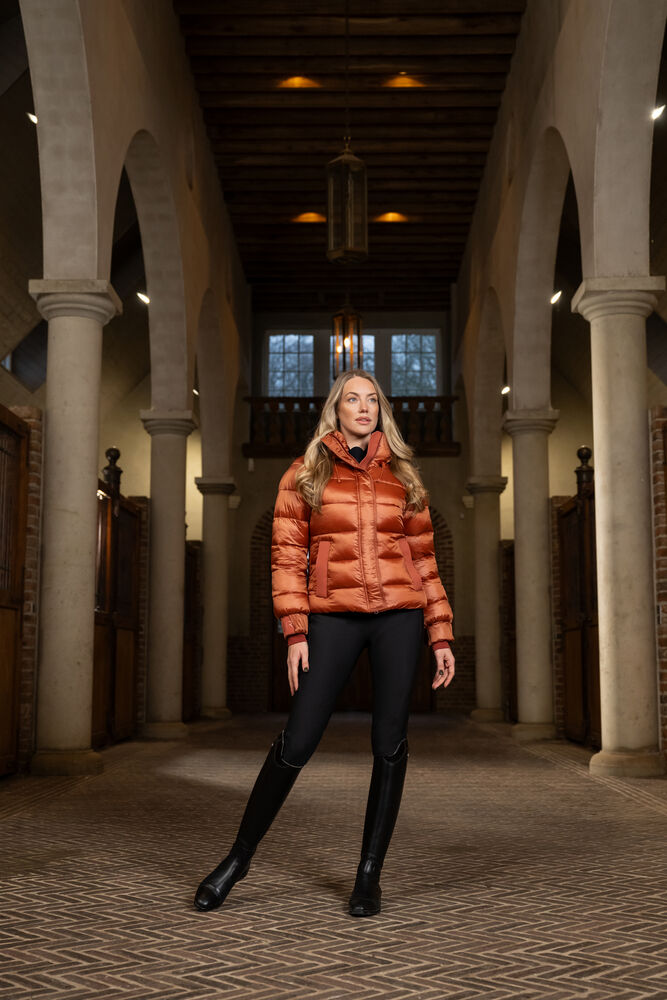Woman wearing an orange puffer jacket in a historic building with arched columns.