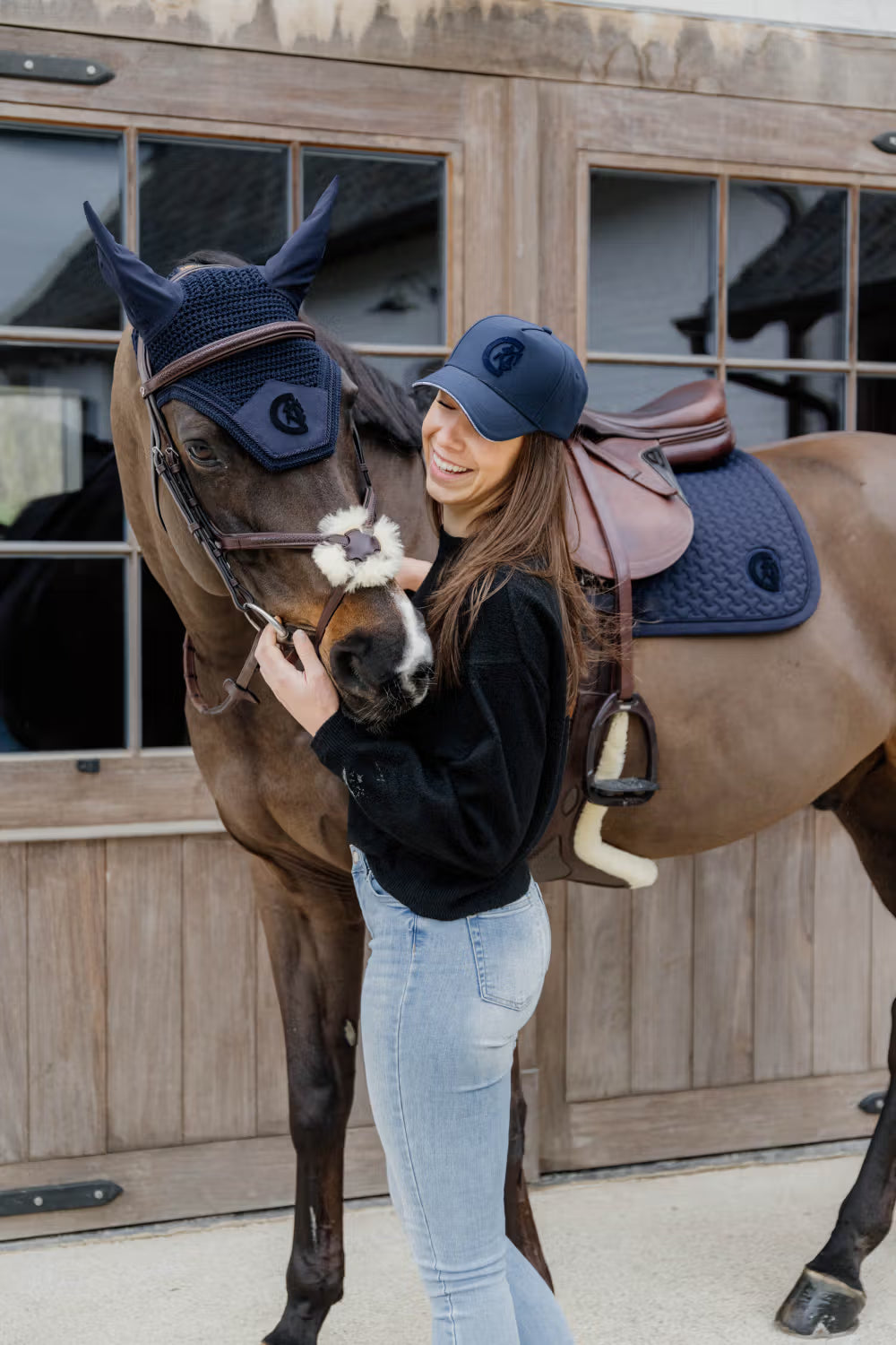 Woman standing next to a horse wearing equestrian gear in front of a wooden stable.