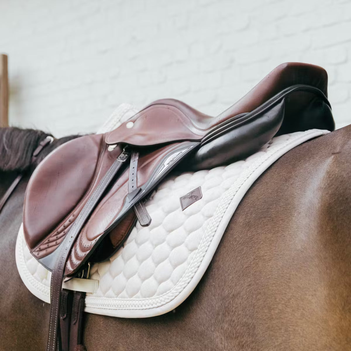 Brown leather saddle on a horse with a beige quilted pad against a light gray background