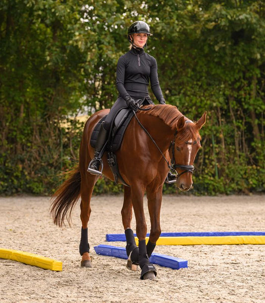 Woman riding a brown horse in an outdoor equestrian setting with greenery in the background.