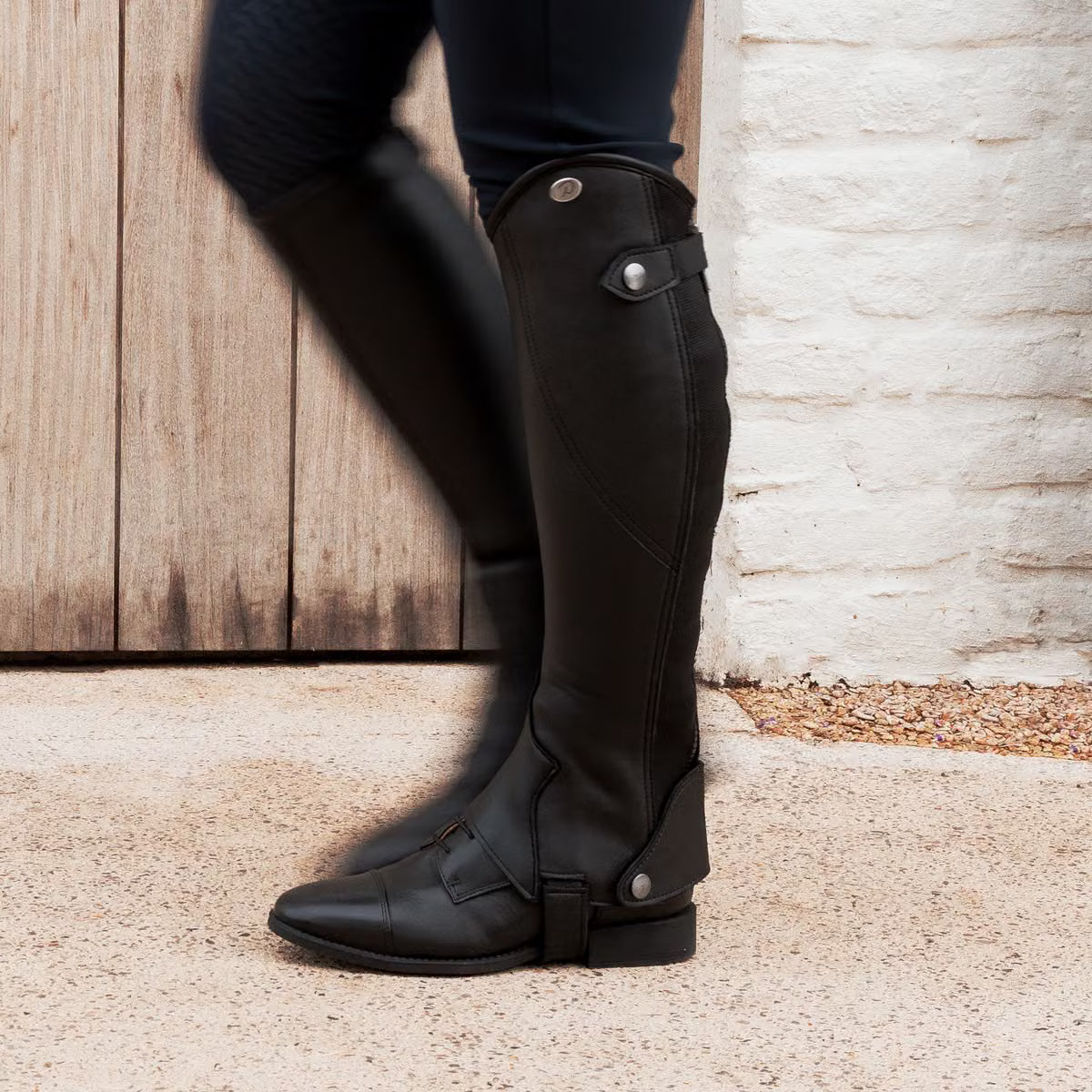 A black leather knee-high riding boot with a pointed toe, low block heel, and decorative strap detail near the top and ankle, shown against a plain white background.
