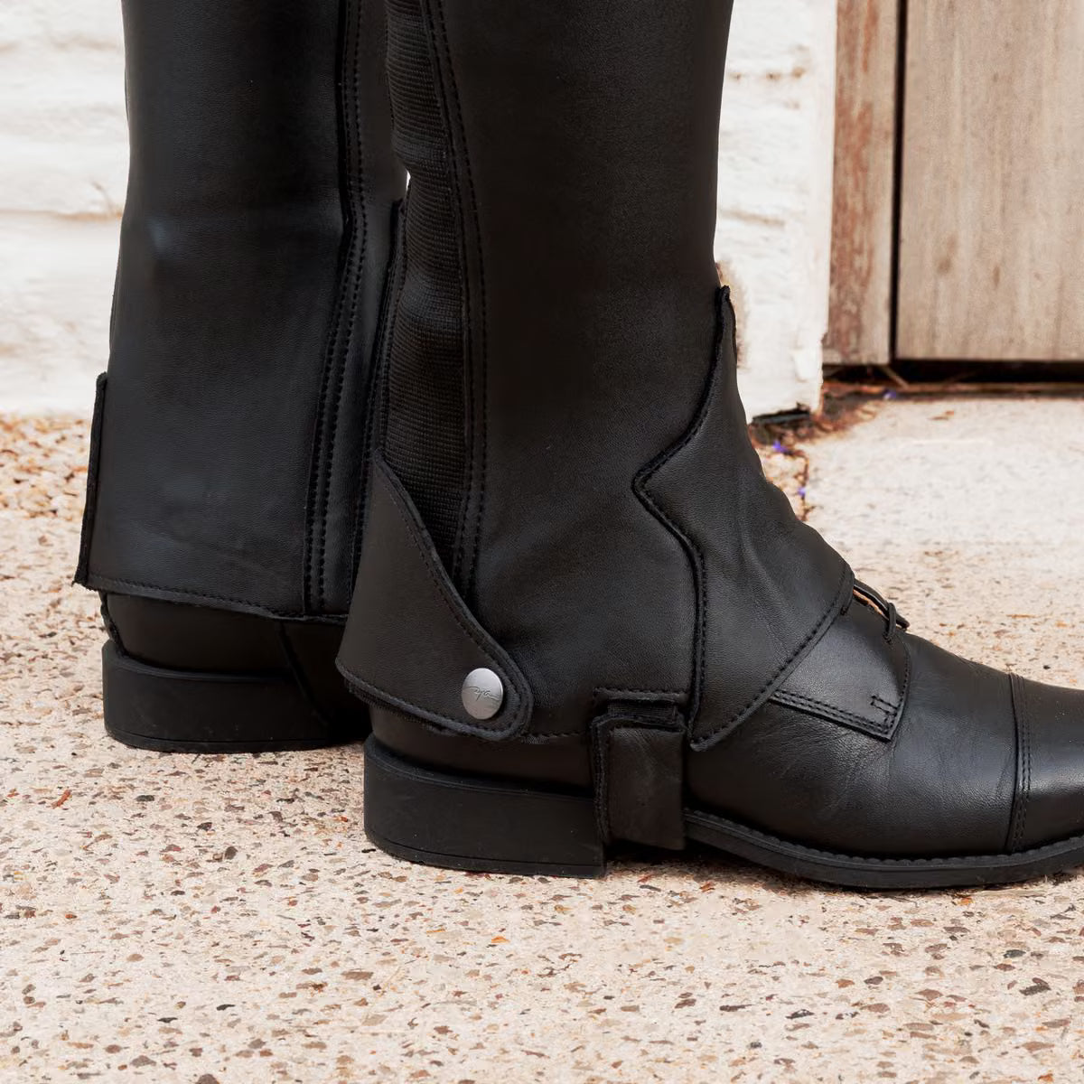 Close-up of a pair of tall black riding boots with ankle straps and a silver snap detail, shown on a person's feet standing on a speckled stone floor.