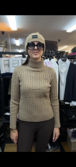 A woman wearing a beige cable-knit turtleneck sweater, a matching knit beanie, and dark sunglasses, standing in a clothing store.