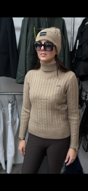 A woman wearing a beige cable-knit turtleneck sweater, matching beanie, and black sunglasses, standing in a clothing store.