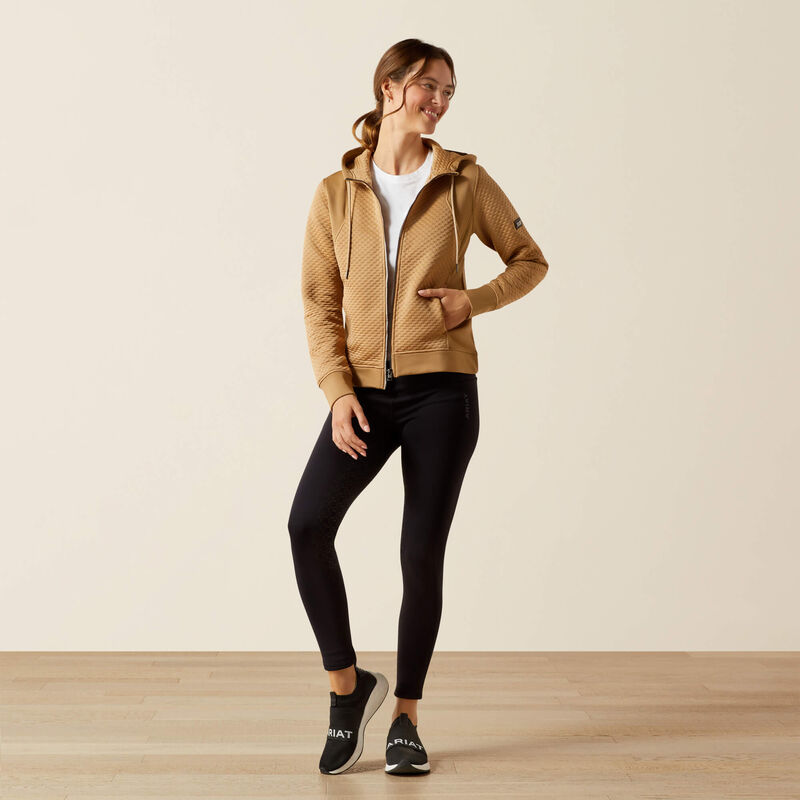 Woman wearing a beige quilted jacket and black leggings on a plain background