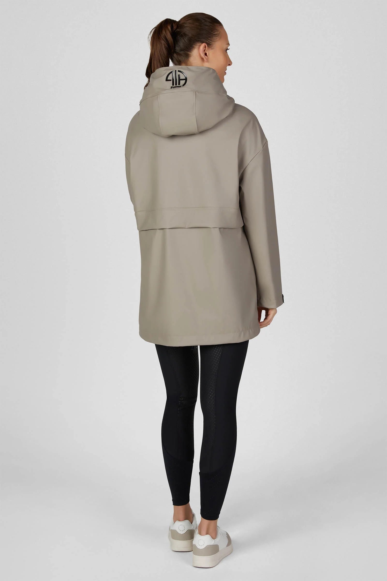 A woman shown from the back wearing a light taupe hooded jacket, black leggings, and white sneakers against a plain light gray studio background.