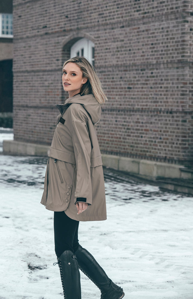 A woman wearing a mid-length beige hooded parka jacket, black pants, and black lace-up boots stands in the snow beside a brick building.