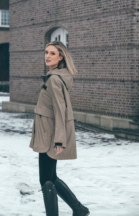 A woman wearing a mid-length beige hooded parka jacket, black pants, and black lace-up boots stands in the snow beside a brick building.