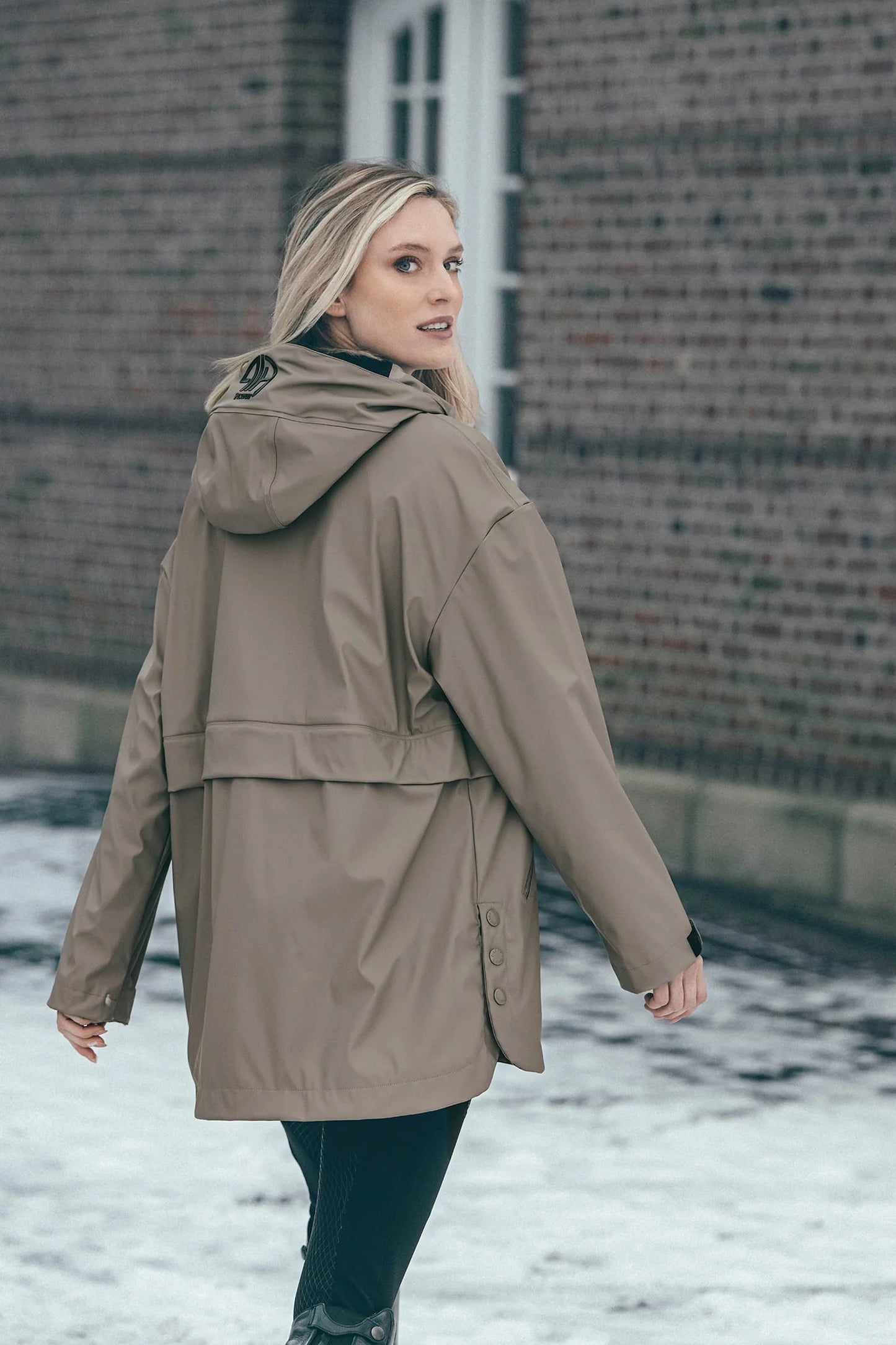 A woman wearing a mid-length hooded raincoat in a taupe-brown color, standing outdoors in front of a brick building with snow on the ground.