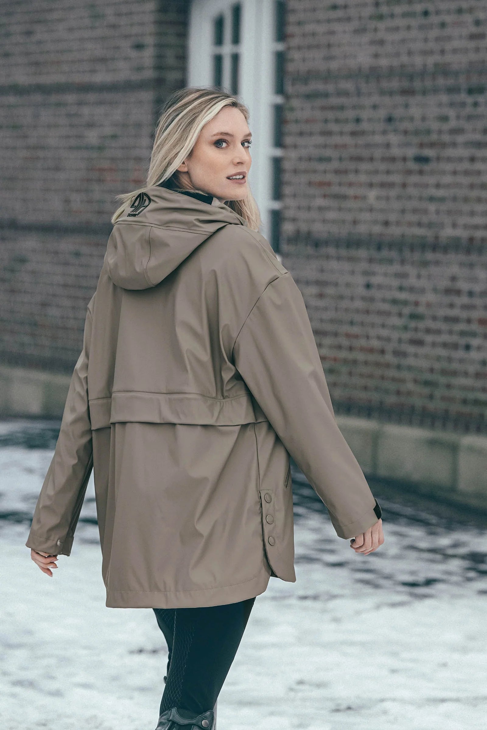 A woman wearing a mid-length hooded raincoat in a taupe-brown color, standing outdoors in front of a brick building with snow on the ground.