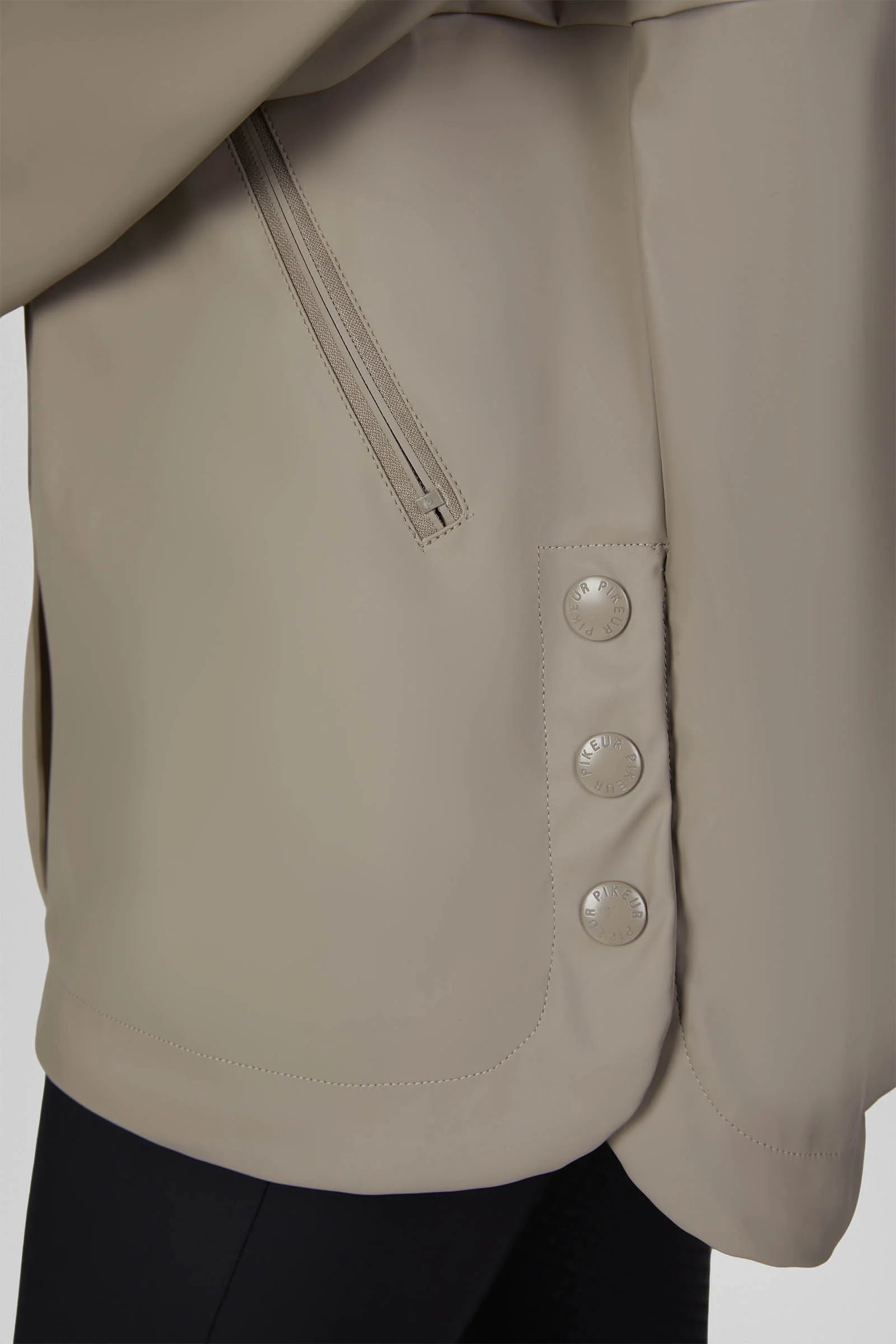 Close-up of a light taupe-beige jacket featuring a diagonal zippered pocket and a vertical pocket with three decorative buttons, worn over dark trousers.