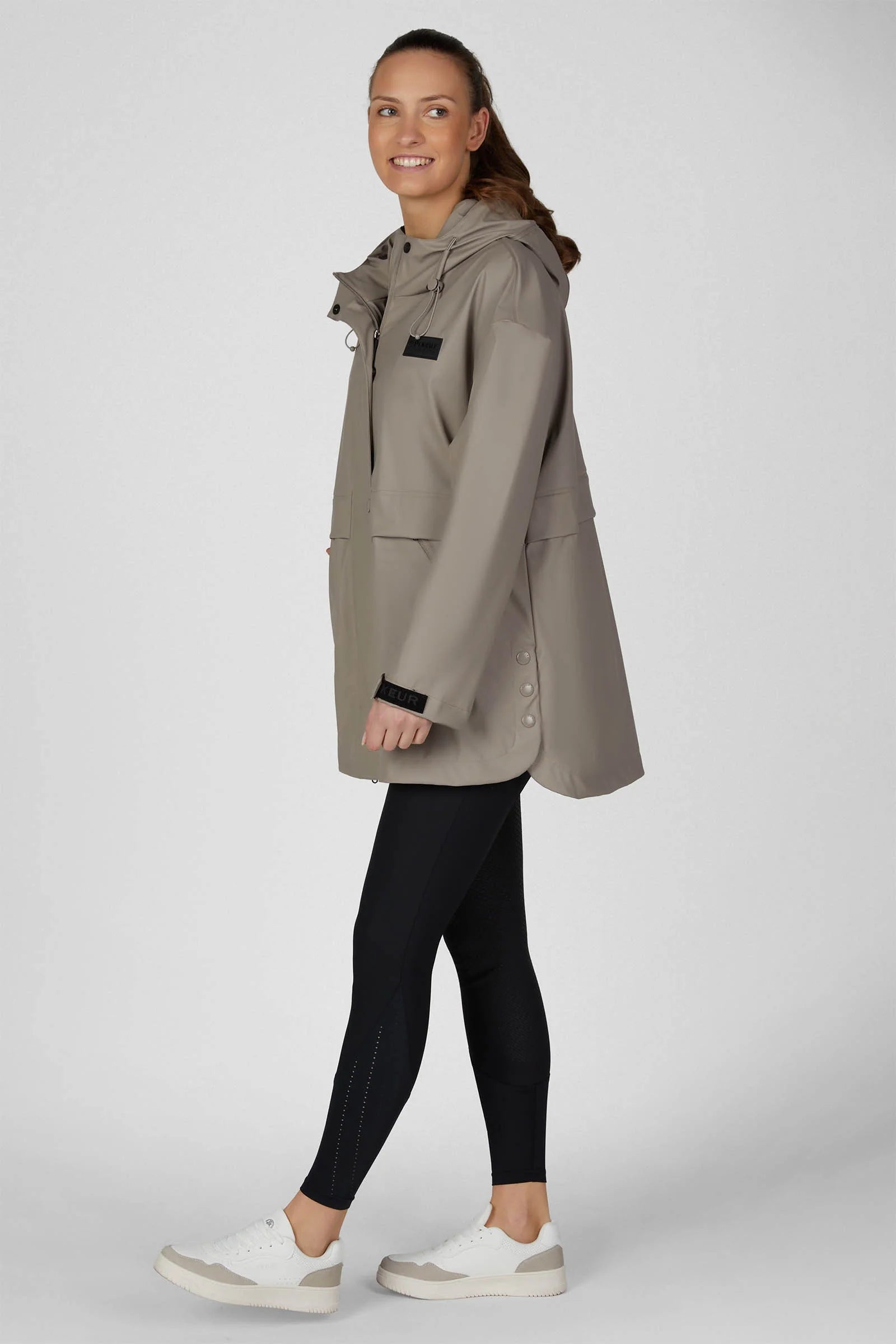 A woman modeling a light taupe hooded jacket, black leggings, and white sneakers against a neutral gray studio background.