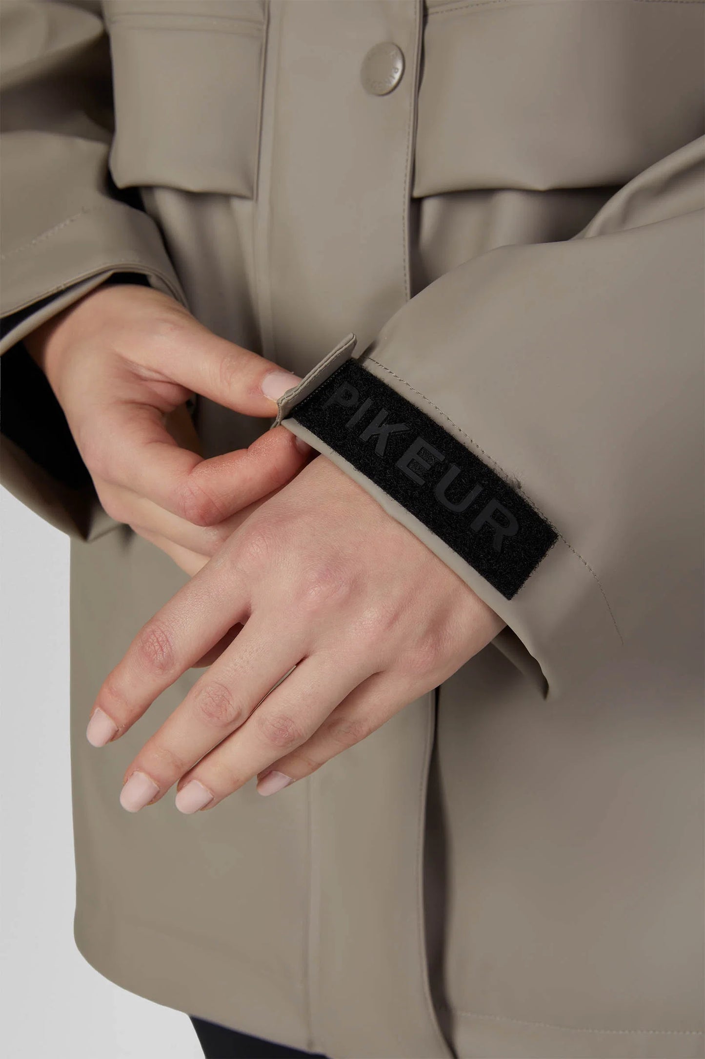 Close-up of a person wearing a beige trench coat and adjusting the sleeve, which features a black hook-and-loop patch with the brand name 'PIKEUR' in beige.