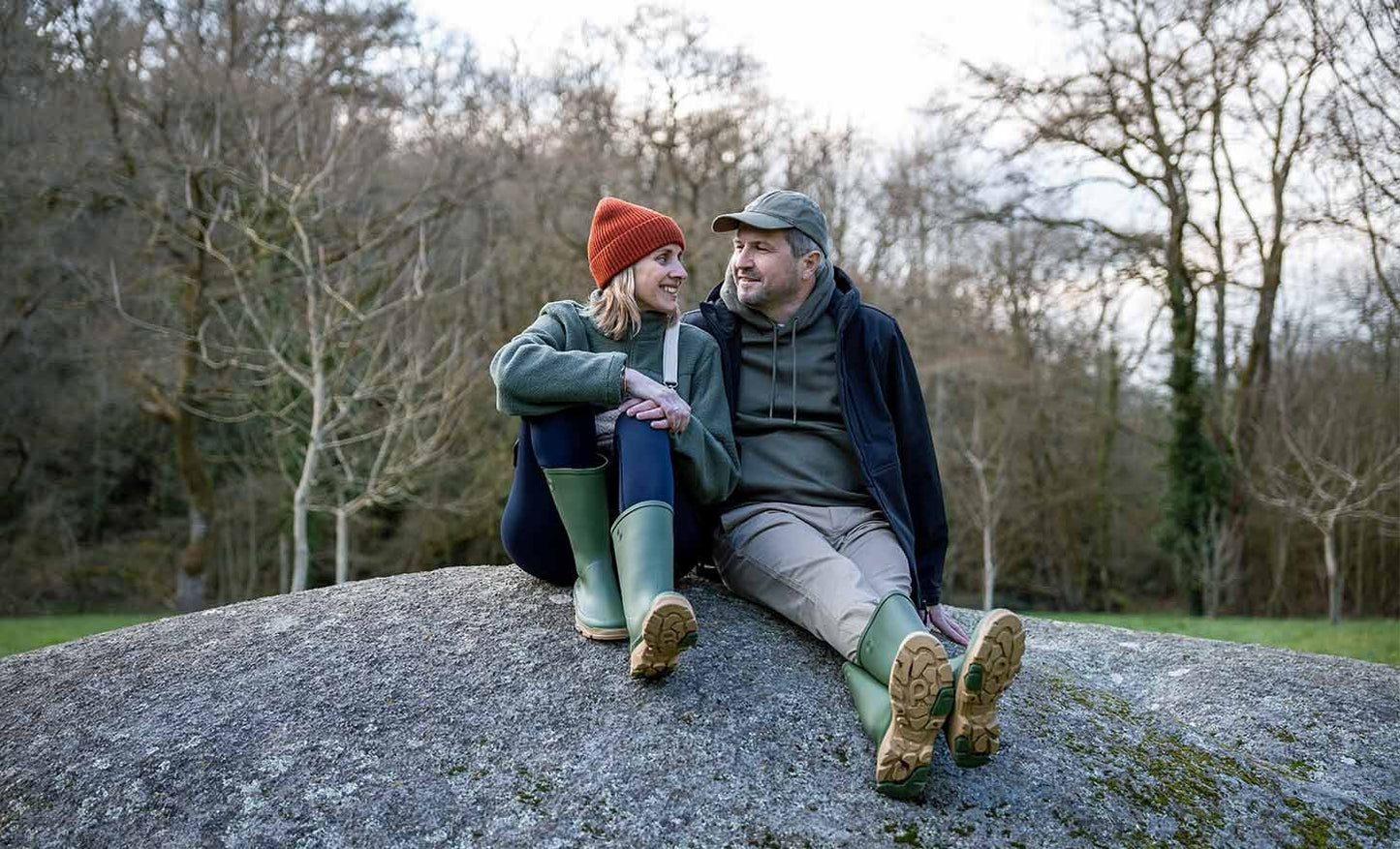 Two people sitting on a rock in a forested area, wearing matching green boots.