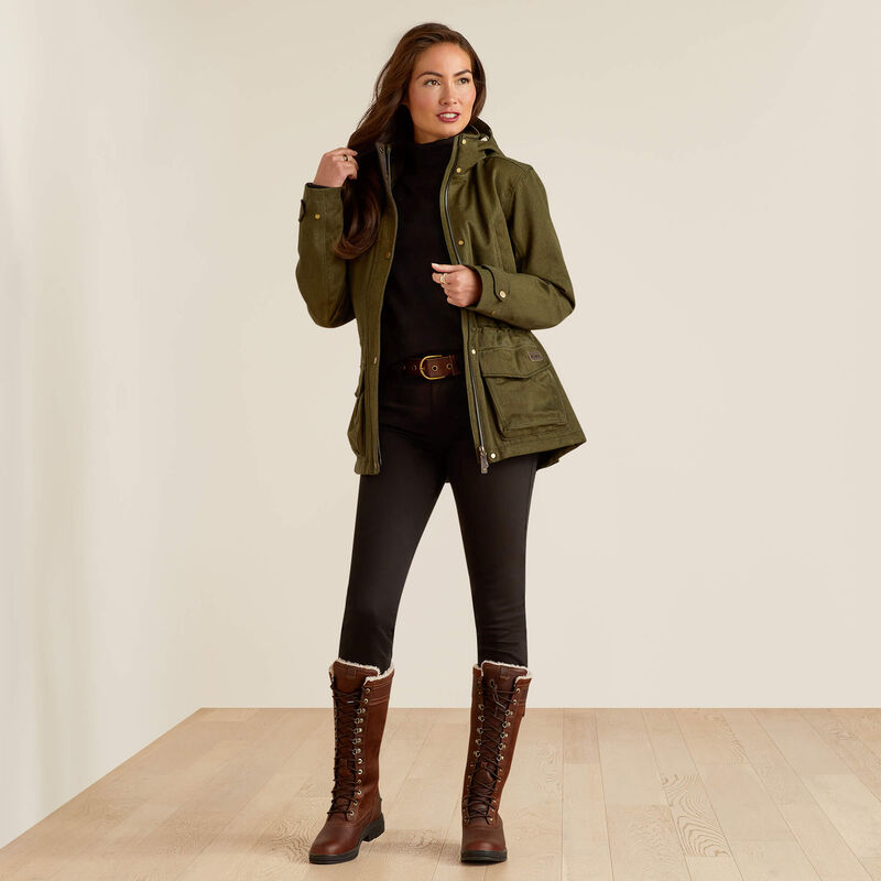 A woman modeling an olive green hooded utility jacket over a black turtleneck, black pants, and brown lace-up boots, standing against a plain beige studio wall on a light wood floor.
