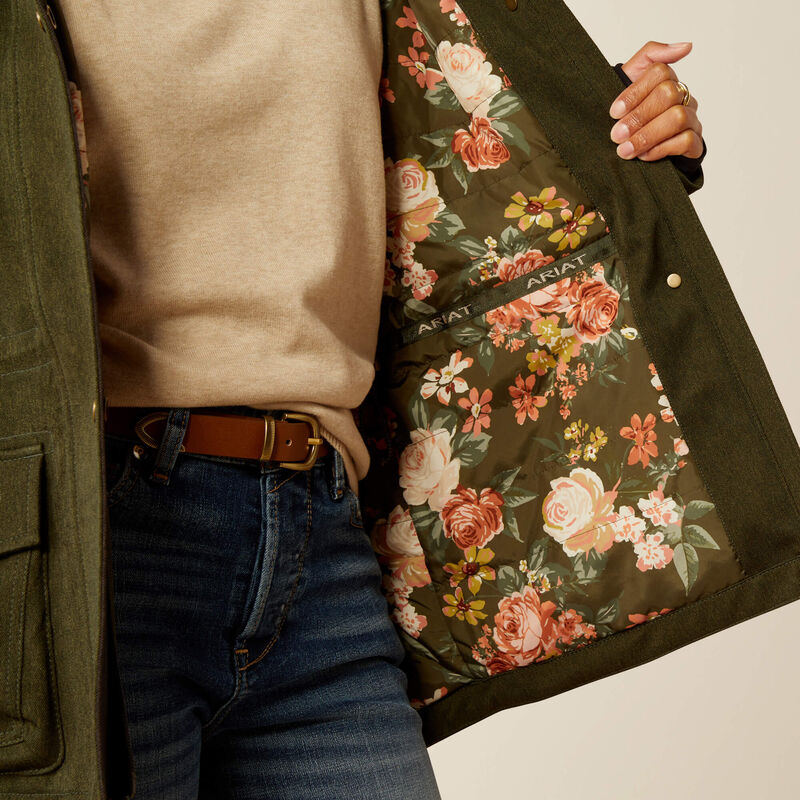 Close-up of a person wearing an olive green jacket with a floral lining, paired with a beige sweater, brown belt, and blue jeans. The hand holding the jacket shows a floral patterned interior with pink, orange, and yellow flowers on a dark green background.
