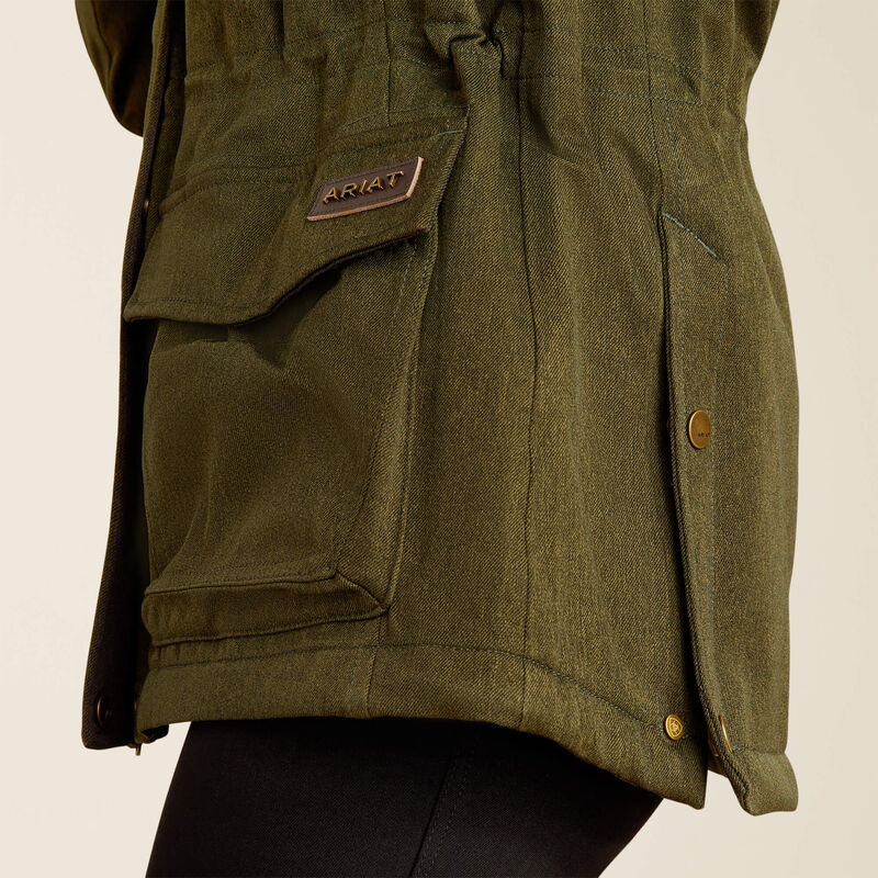 Close-up of an olive green Ariat jacket with a flap pocket, metal brand patch, and snap-button closure, worn over black pants against a light neutral background.