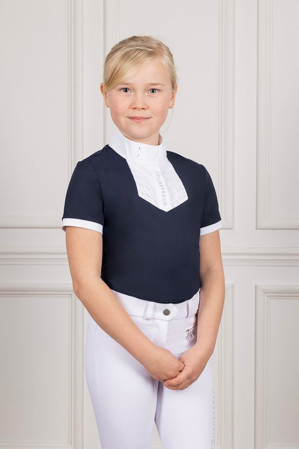 Young girl wearing a navy equestrian shirt with white trim and white pants against a light-colored wall.