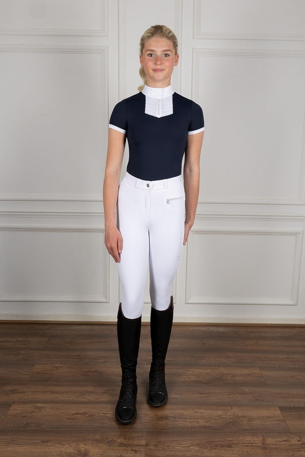 Person wearing equestrian attire including a navy top, white pants, and black boots against a neutral background.