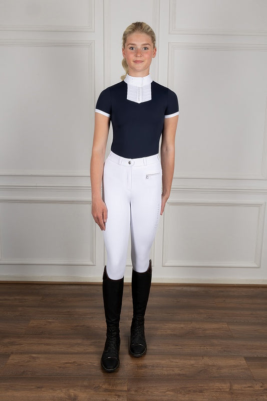 Person wearing equestrian attire including a navy top, white pants, and black boots against a neutral background.