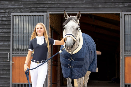 Woman in equestrian attire standing next to a horse wearing a blue rug in front of a stable.