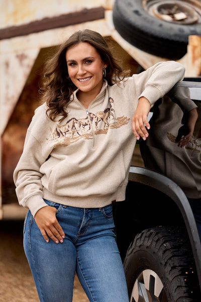 A woman wearing a beige pullover hoodie with a brown graphic print of horses and a rider, paired with blue jeans, posing next to a vehicle in an outdoor rustic setting.