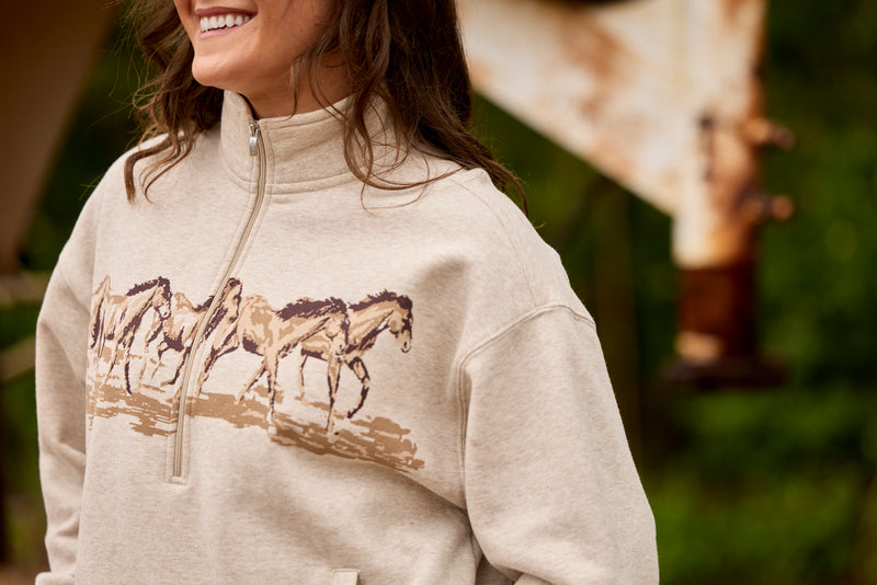 A woman wearing a light beige quarter-zip sweatshirt with a graphic print of four horses in brown and tan, set against a blurred outdoor background.