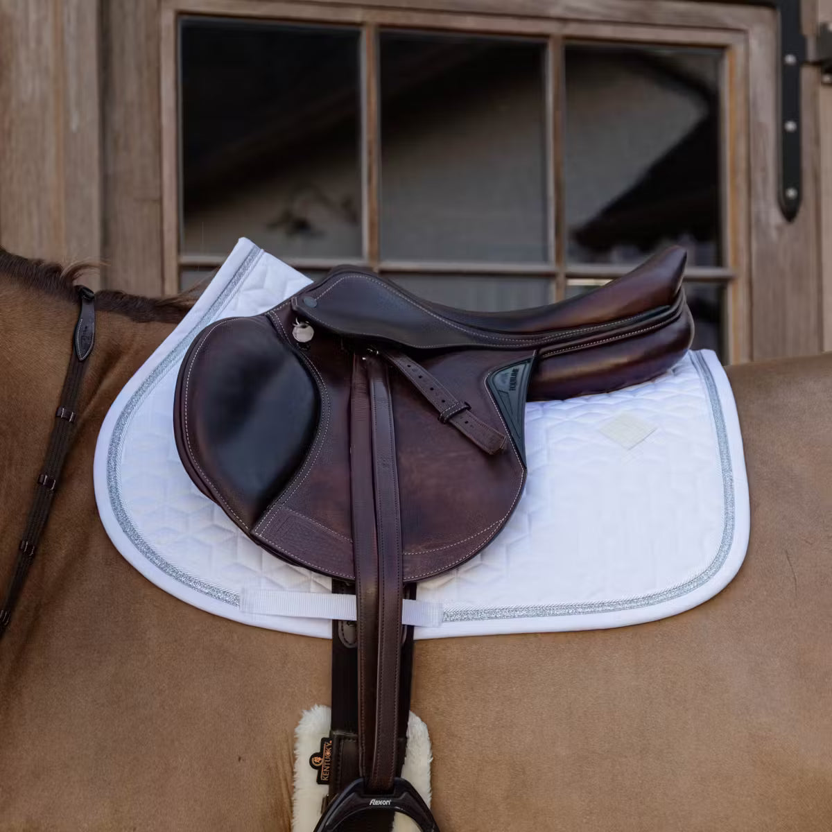 Brown saddle on a horse with a white saddle pad, wooden stable in the background