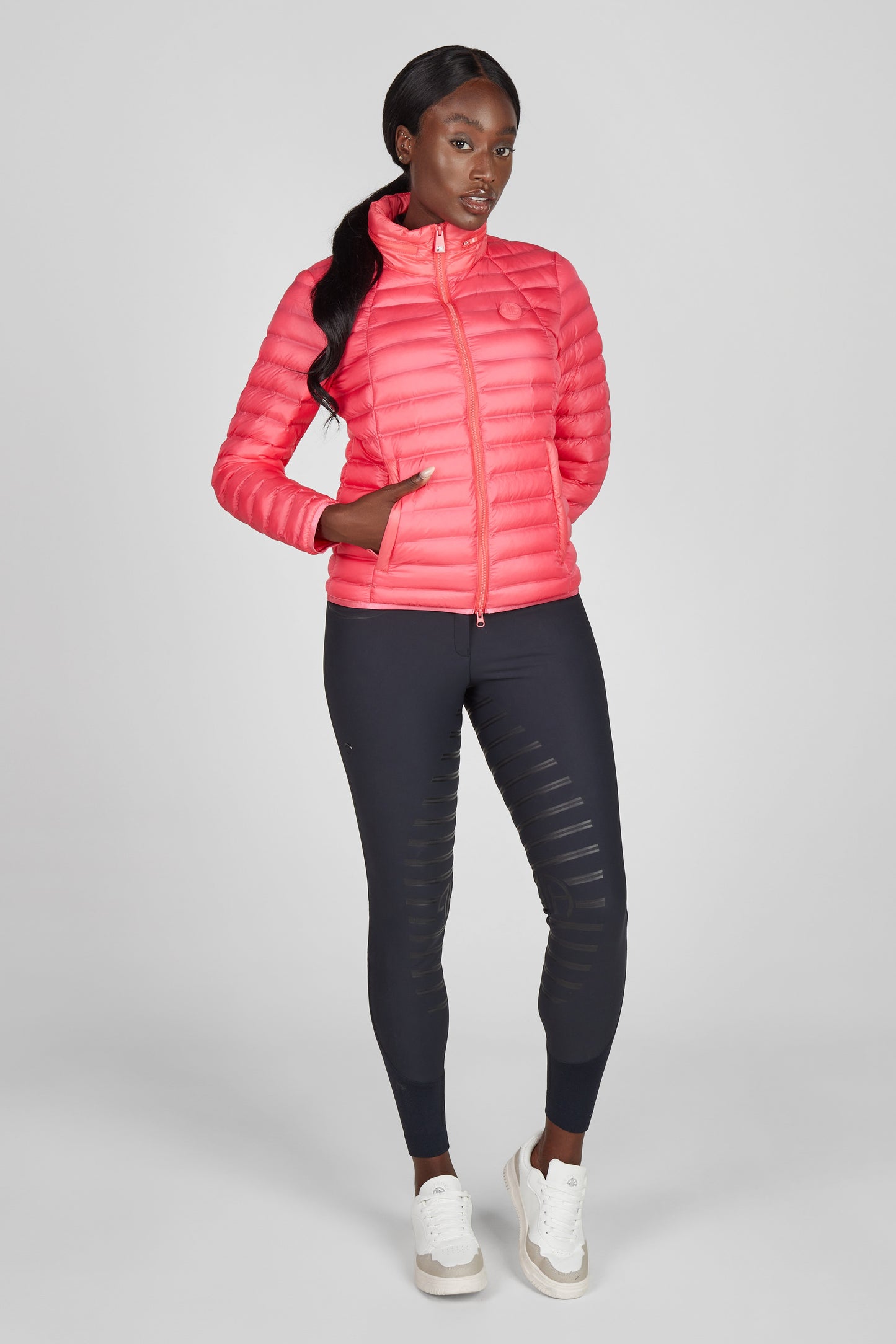 Model wearing a pink puffer jacket and navy breeches on a grey background
